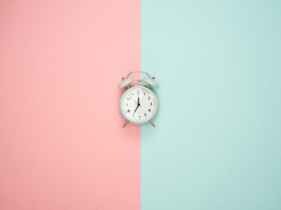 Ao centro da imagem um relógio. De fundo, metade em rosa, metade em verde, ambos em tons pastéis.
